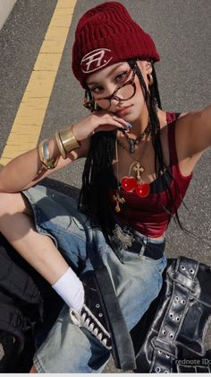 Person in a red beanie and denim shorts with a cherry necklace sitting on asphalt