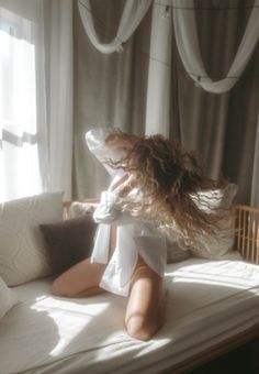 Woman with long curly hair kneeling on a bed in a white shirt