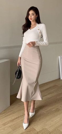 Woman in a white top and mermaid skirt holding a black handbag indoors