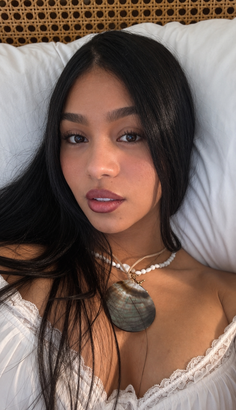 Woman with long black hair wearing a white top and shell necklace, lying on a pillow