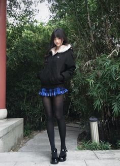 Woman in a black jacket and blue plaid skirt standing outdoors among bamboo