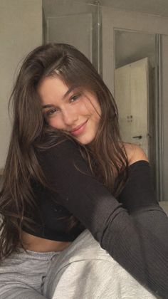 Smiling woman with long brown hair wearing a black off-shoulder top and grey pants indoors