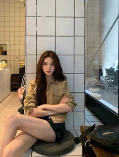 Woman with long hair wearing a tan jacket and black shorts, sitting indoors