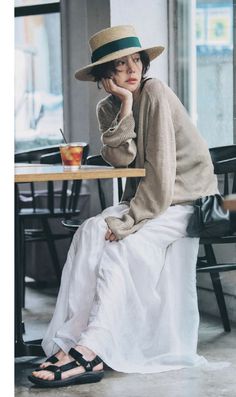 Woman in a straw hat, sweater, and long white skirt sitting at a table indoors