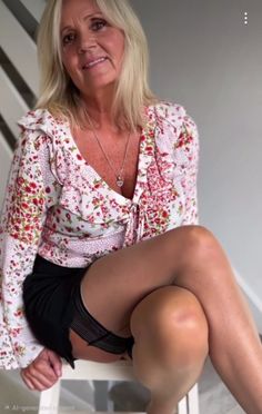 Woman with blonde hair in a floral blouse and black shorts, sitting indoors