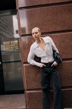 Woman with blonde hair wearing a cardigan and black jeans leaning against a building