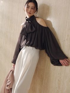 Woman in a dark off-shoulder top with puffy sleeves and white pants leaning against a wall