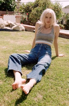 Woman with blonde hair in a crop top and jeans sitting on grass outdoors