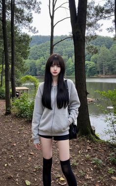Woman with long black hair wearing a grey hoodie and black shorts by a forest lake