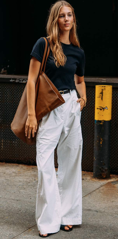 Woman with long hair wearing a black t-shirt and white wide-leg pants with a brown tote bag