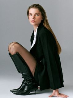Woman in a black blazer, white shirt, and black boots posing in a studio