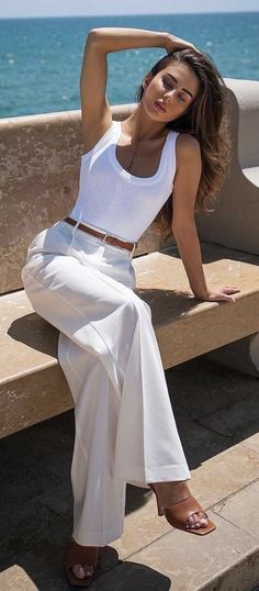 Woman in a white tank top and white pants sitting outdoors with the ocean in the background