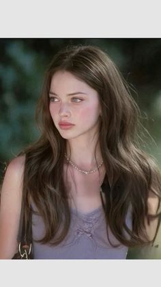 Woman with long brown hair and a pearl necklace wearing a lavender top outdoors