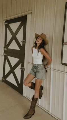 Woman in a cowboy hat, denim shorts, and cowboy boots leaning against a wall