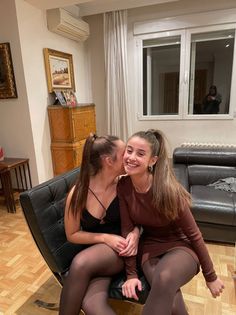 Two young women with ponytails sitting together indoors, one whispering to the other