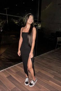 Woman in a black dress with a high slit and sneakers, smiling indoors at night