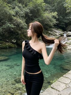 Woman in a black one-shoulder top and jeans by a clear stream in a forest