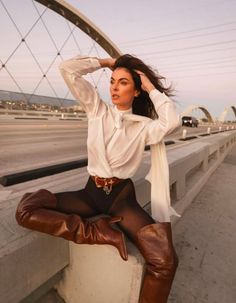 Woman in a white blouse and brown boots posing on a bridge at dusk