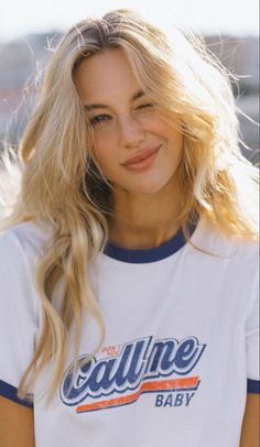 Blonde woman winking in a white t-shirt with text, outdoors with soft lighting