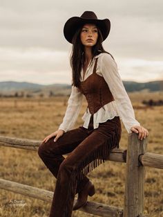 Woman in a cowboy hat and suede outfit sitting on a wooden fence in the countryside