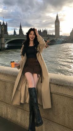 Woman in a trench coat and mini skirt sitting by a river with a cityscape background