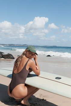 Woman in a dark swimsuit and green cap squatting next to a surfboard on a beach