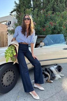 Woman in a striped shirt and jeans with a dog next to a convertible car