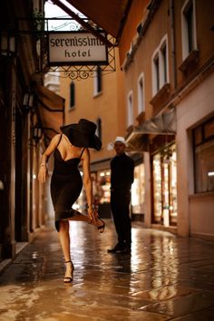 Woman in a black dress and hat adjusting her shoe on a wet street