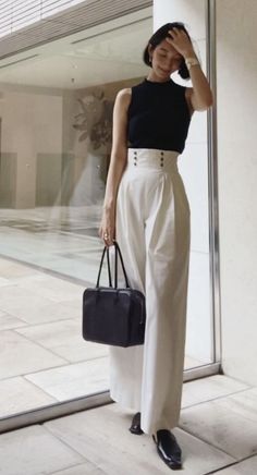 Woman with short hair in a black top and white high-waisted pants holding a black handbag