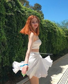 Woman with red hair in a white top and skirt outdoors with a green hedge