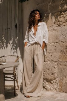 Woman in a white shirt and wide leg pants leaning against a stone wall outdoors