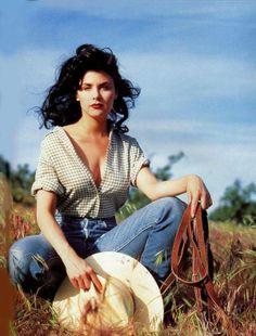 Woman in a gingham shirt and jeans holding a cowboy hat in a field