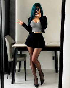 Woman with teal hair in a crop top and pleated skirt posing in a mirror