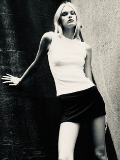 Woman in a white tank top and black mini skirt leaning against a wall