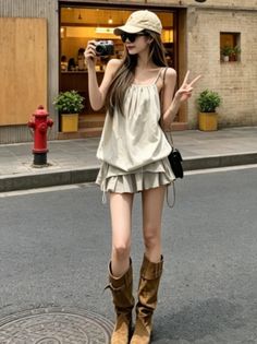 Woman in a mini skirt, boots, and cap holding a camera on a street