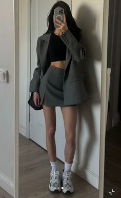 Woman in a gray blazer, mini skirt, and crop top taking a mirror selfie
