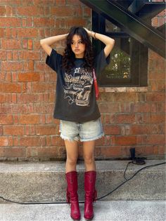 Woman with long hair wearing a graphic t-shirt, denim shorts, and red boots against a brick wall