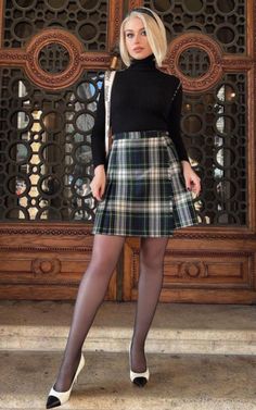 Woman in a plaid skirt and black turtleneck standing in front of an ornate wooden door