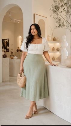 Woman in a white top and green midi skirt holding a straw bag indoors