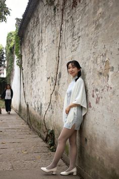 Woman in a light blue cheongsam dress and white shawl leaning against an old wall