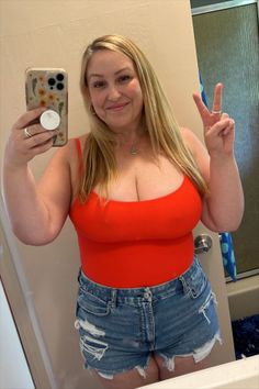Woman with blonde hair in a red top and denim shorts taking a mirror selfie