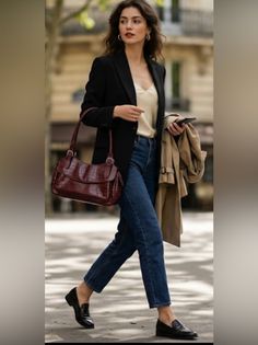 Woman in a black blazer and blue jeans walking outdoors with a shoulder bag