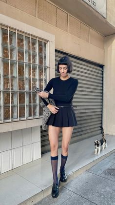 Woman with short black hair in a pleated skirt and knee socks on a street with a cat