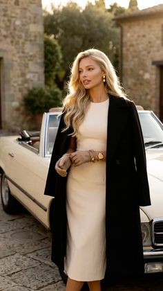 Woman in a black coat and cream dress standing next to a vintage car at golden hour