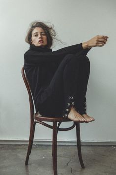 Woman in a black turtleneck and pants sitting barefoot on a wooden chair