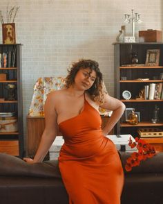 Woman in an orange one-shoulder dress leaning on a couch indoors