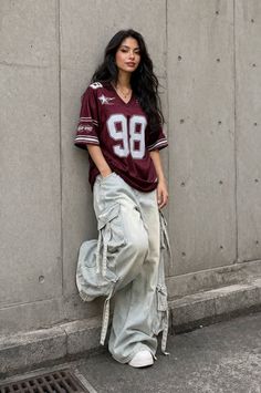 Woman with long dark hair wearing a jersey shirt and cargo pants leaning against a wall