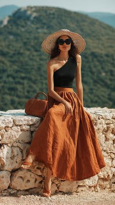 Woman in a straw hat and long skirt sitting on a stone wall with a mountain view