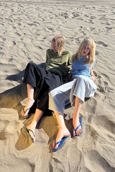 Two people with blonde hair sitting on a sandy beach in flip flops