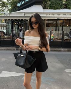 Woman in an off-shoulder top and shorts holding a handbag on a street in front of a cafe